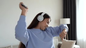 Happy young teenage girl in cornflower blue sweater dancing at home having fun listening to music wearing headphones enjoying weekend celebration with funky dance moves. 4k footage