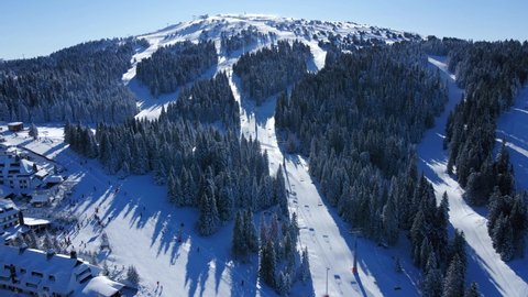 Aerial view of famous Kopaonik ski resort town in winter, buildings and slopes covered in snow, clear blue sky - landscape panorama of  mountain range from above, Serbia.