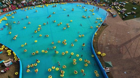 Crowd of people relaxes in the city open-air water park, people swim in a large pool on inflatable rings and sunbathe on sun loungers.