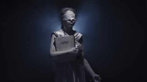 Greek goddess of justice stands on a black background with moving smoke. Blindfolded woman holds book of justice in her hand and lifts the scales. Studio. Concept of justice.