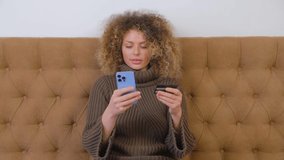 Happy buyer woman paying with credit card in smartphone app. Cheerful white female using modern blue mobile phone for online shopping at home. Beautiful person with curly hair buying in web store