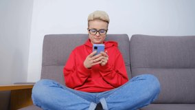 White woman typing comment online on social media app in smartphone. Cheerful tom boy female with dyed short hair typing message on modern mobile phone while sitting on couch at home during lockdown