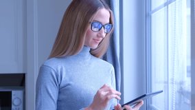 Young woman using tablet pc near window at home. Beautiful blonde female in glasses browsing internet news feed on modern mobile gadget in 4k video. Online communication and entertainment concept