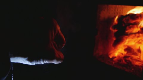 A man warms his hands after hypothermia while sitting by a warm hearth in the dark. Water vapor escapes from the skin. Extreme living conditions