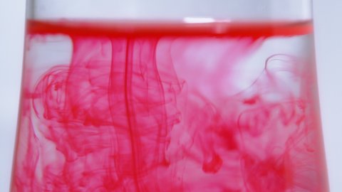 Pink color ink drips and drops flow in an abstract water fluid. Splashes and splatter of bright red paint tint. Texture, patterns and trails of rosy dye flowing in glass of pure transparent liquid.