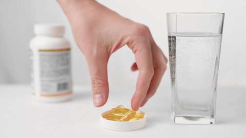 A woman's hand takes a capsule of organic vitamins from the table and drinks it down with a glass of water. Taking vitamin E, fish oil, Omega 3, vitamin D. Taking dietary supplements.