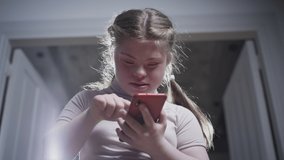 A girl with down syndrome is holding a smartphone. Writes a message, looks at photos, uses a social media app. Disabled person at home. Life with a disability. - Powered by Shutterstock - Get 15% off with code: PIKWIZARD15