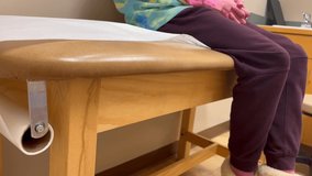 Young kid child girl or boy with covid-19 at Doctor’s office hospital visit waiting on examination exam table with paper roll nervously fidgeting kicking feet wide table focused clip.