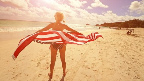 backside of Hawaiian woman holding at sunset an American flag wearing in American flagged bikini. Tropical Lanikai Beach, east shore of Oahu in Hawaii, USA. Freedom and 4th July patriotic concept.