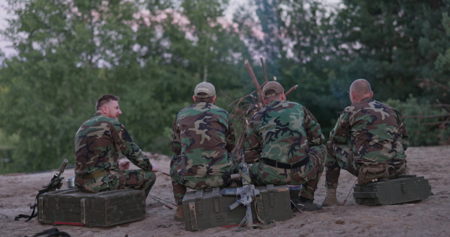 Hunters in camouflage clothing after hunting game light a bonfire, fry sausages, tell each other a story, view from behind, hidden weapons, rest, smoke cigarettes