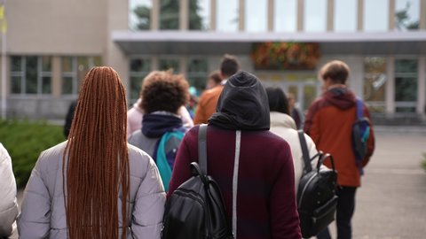 Rear view of multi-ethnic secondary school pupils with backpacks walking to school building for lessons. Group of multiracial teenage schoolkids outdoor on the way to high school