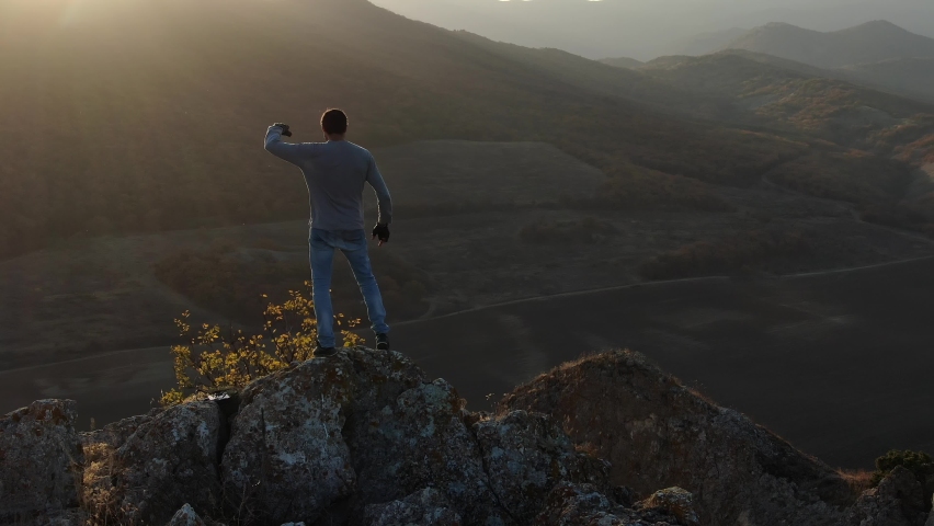 A man rejoices in life standing on top of a cliff against a backdrop of mountain slopes and valleys with vineyards and a village in autumn