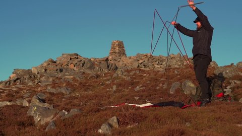 A man setting up a camping tent on a mountain summit at sunset