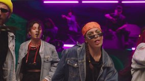 Group of stylish young people dancing , moving near military tank inside garage building Colorful stylish clothes , hippie style fashion . Modern dances . Purple led light , slow motion