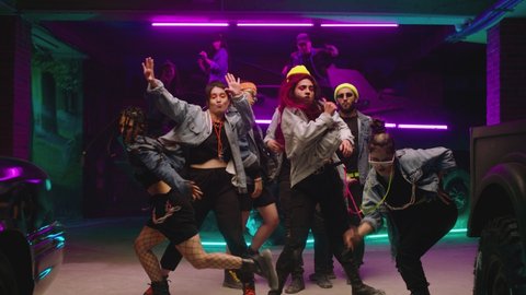 Group of stylish young people dancing , moving near military tank inside garage building Colorful stylish clothes , hippie style fashion . Modern dances . Purple led light , slow motion Adlı Stok Video