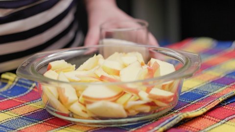 senior woman pouring sugar in glass bowl with chopped apples during making apple pie at domestic kitchen