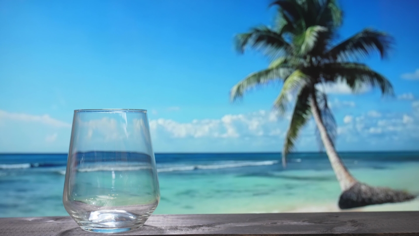 a fizzy cola drink with ice cubes is poured into the glass. A cold soda against the backdrop of a beach with a palm tree, a cocktail with moving bubbles, a summer party on the beach.