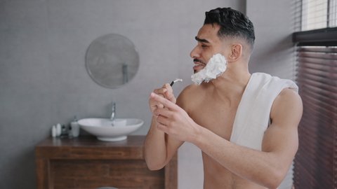 Cheerful funny goofy handsome arab man guy with white soap suds on beard naked male with towel on shoulder singing song into razor having fun shaving in bathroom at home dancing music humorous hygiene