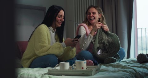 Cinematic shot of young girlfriends or sisters are choosing with smartphone design of warm clothes to be knitted with yarn wool at home during covid-19 pandemic lockdown social distancing.