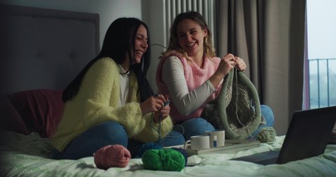 Cinematic shot of young creative girlfriends or sisters knit together warm yarn wool with knitting needles while watching online tutorial with laptop on bed in room at home during covid-19 pandemic.