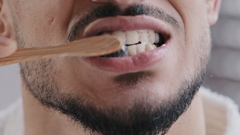 Extreme close-up unrecognizable bearded man brushing teeth with white paste whitening agent with wooden toothbrush clean oral dental health at home bathroom morning routine daily hygiene care concept