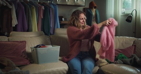 Cinematic shot of young happy creative woman tries softness of sweater of warm yarn wool she finished to knit with knitting needles while sitting comfortably on sofa in living room at home.