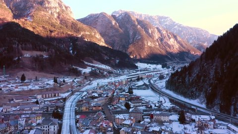 Drone going forward creating a beautiful panorama of the Pontebba city in Udine. Beautiful aerial perspective. unrise with warm colours. In background Italian Alps. Winter landscape with snow