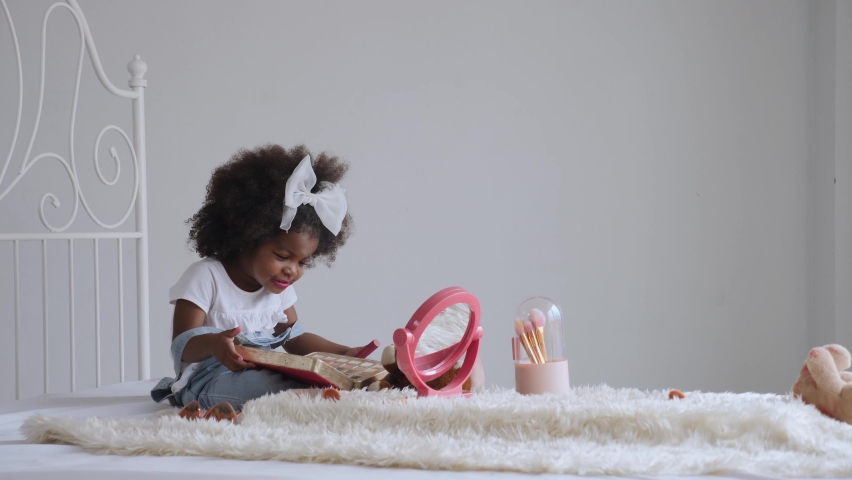 Cute African preschool girl with afro hair sitting on bed, playing alone, applying make up, looking at the mirror, having fun, smiling, making messy face. Naughty and smart child acitivity concept Royalty-Free Stock Footage #1086460760