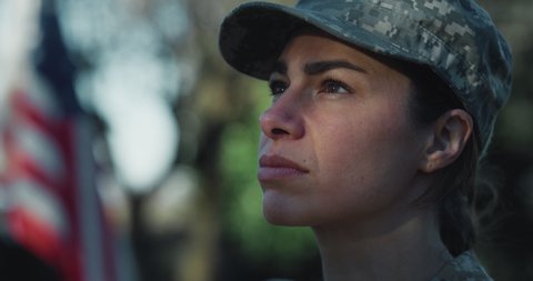 Cinematic close up shot of serious determined responsible female patriot soldier in camouflage military uniform is looking around outdoors with american flag on background.