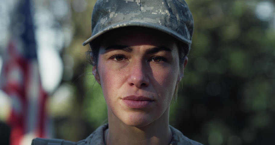 Cinematic close up shot of serious determined responsible female patriot soldier in camouflage military uniform is looking in camera outdoors with american flag on background.