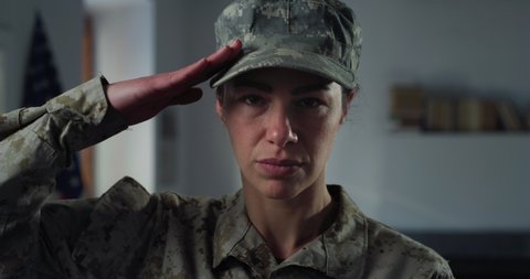 Cinematic close up shot of serious determined responsible female patriot soldier in camouflage military uniform and cap saluting and looking at camera indoors with american flag on background.