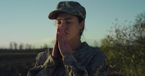 Cinematic close up shot of young serious determined responsible female patriot soldier in camouflage military uniform and cap is praying with hope on fields at sunset.