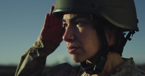 Cinematic close up shot of young serious determined responsible female patriot soldier in camouflage military uniform and helmet is saluting and looking on horizon on fields at sunset.