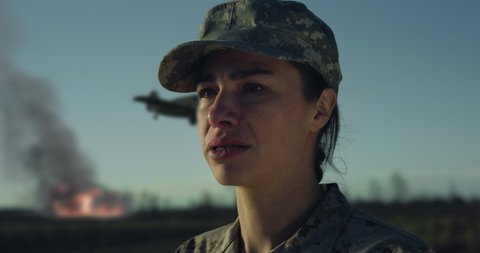 Cinematic close up shot of young suffering desperate despondent female patriot soldier in camouflage military uniform and cap is crying with emotions of sorrow during operation on fields at sunset.