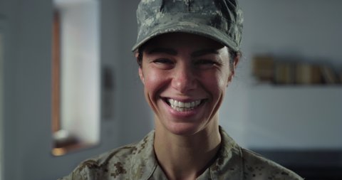 Cinematic close up shot of young serious determined responsible female patriot soldier in camouflage military uniform and cap looking and smiling in camera indoors.