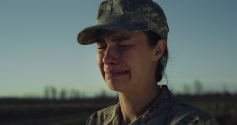 Cinematic close up shot of young suffering desperate despondent female patriot soldier in camouflage military uniform and cap crying with emotions of sorrow after operation on fields at sunset.