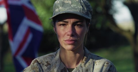 Cinematic close up shot of young serious determined responsible female patriot soldier in camouflage military uniform is looking in camera outdoors with british flag on background.