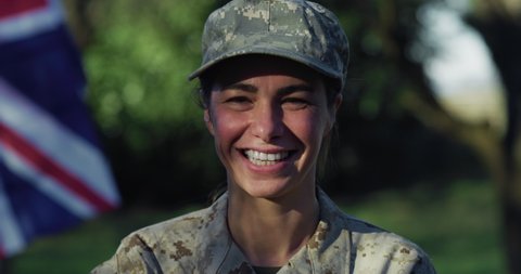 Cinematic close up shot of young determined responsible female patriot soldier in camouflage military uniform and cap is looking and smiling in camera outdoors with british flag on background.