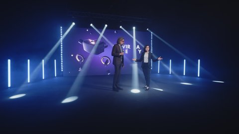 A female presenter interviewing a male developer of a new augmented reality device during the presentation of meta universe in a modern studio with spotlights, near the LED screen in 3D objects