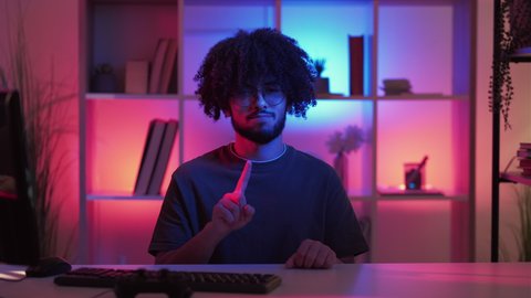No gesture. Skeptic man. Meme expression. Disagreed smiling hipster guy waving finger sitting desk with computer in dark neon light home interior looped.