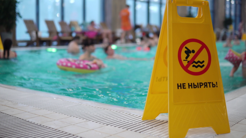 The inscription in Russian do not dive. A yellow sign stands near the pool with a warning inscription against the background of people floating in the pool 4k | Shutterstock HD Video #1086475253