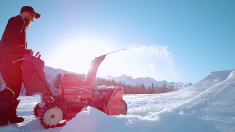 SLOW MOTION, COPY SPACE, LENS FLARE, CLOSE UP: Bearded man operates a snow blower on a sunny winter day. Male ski resort maintenance worker uses a snowblower on a picturesque sunny December day.