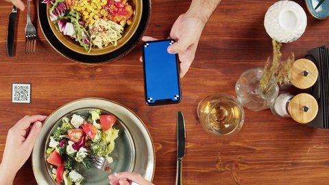 Woman using smartphone with blue screen and eating poke bowl salad, qr code with online menu in cafe. Modern technology, ordering meal in application. Contactless touch-free payment in app.