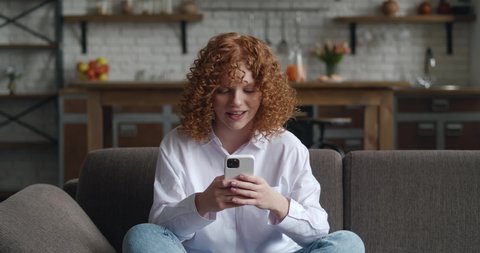 Overjoyed young redhead woman using smart phone winning gift or prize in social media app sit on sofa at home. Excited happy woman winner celebrating mobile victory at living room, showing yes gesture