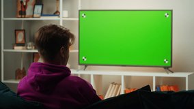 Man watching television in living room. Young guy sitting on sofa against tv with green screen, back view. Male person using smartphone with chroma key.