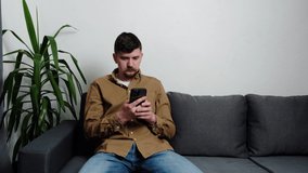 Handsome young man relaxing on comfortable grey couch at home, using mobile application in living room. Bearded caucasian guy chatting in social network, dating online or web surfing internet