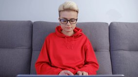 Young woman using laptop computer on couch in living room. Tomboy person with short dyed hair typing text on notebook pc. Professional freelancer female doing distant work online during lockdown