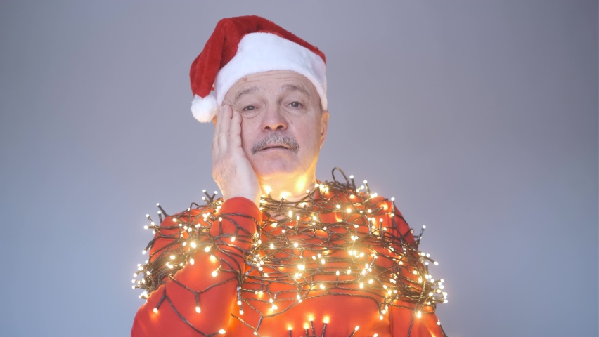 Senior sad man in red christmas hat wrapped in garland.