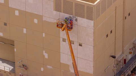 Construction workers in bucket of crane vehicle for restore and repair of the facade of building aerial timelapse. Painting the wall in gray color