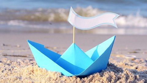 Blue paper boat on a sandy beach near the sea on a sunny summer day. In boat stick with space for text. Small boat from paper on sand near sea waves. Concept Travel, tourism, vacation, rest, holilday